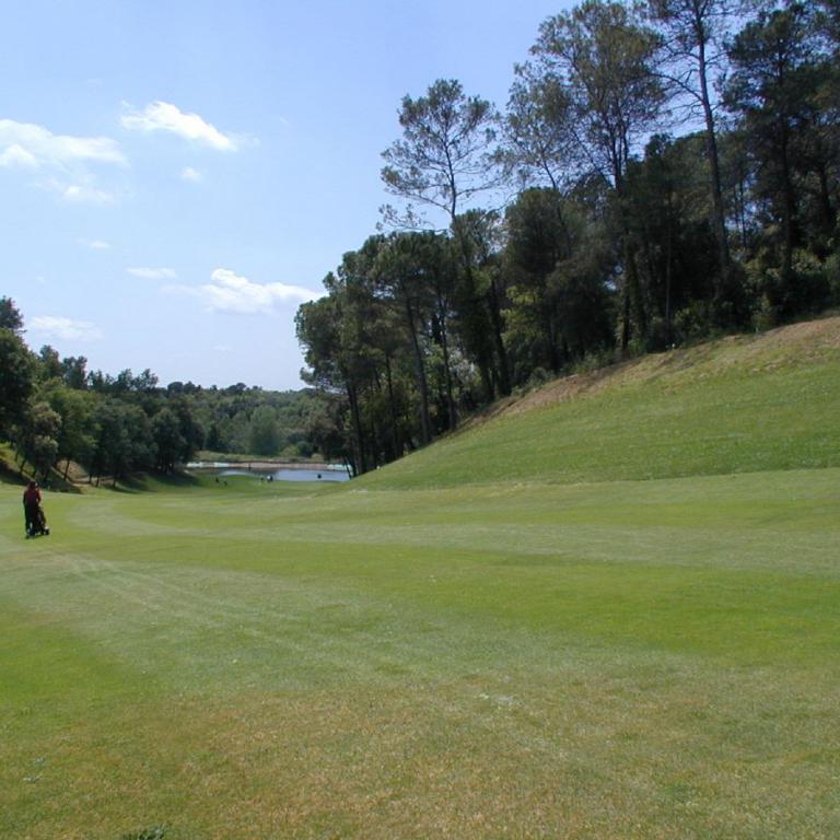 Eine weite grüne Golfplatzlandschaft, die zu einem bewaldeten Hang abfällt, unter einem hellblauen Himmel.