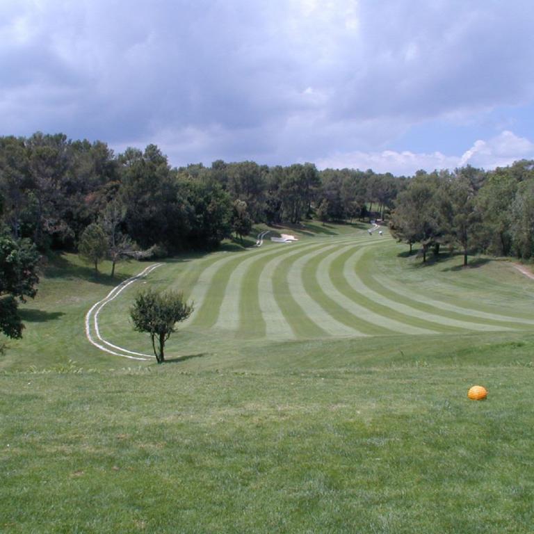 Ein weitläufiger Golfplatz mit gestreiftem Fairway und umliegenden Waldgebieten unter einem bewölkten Himmel.