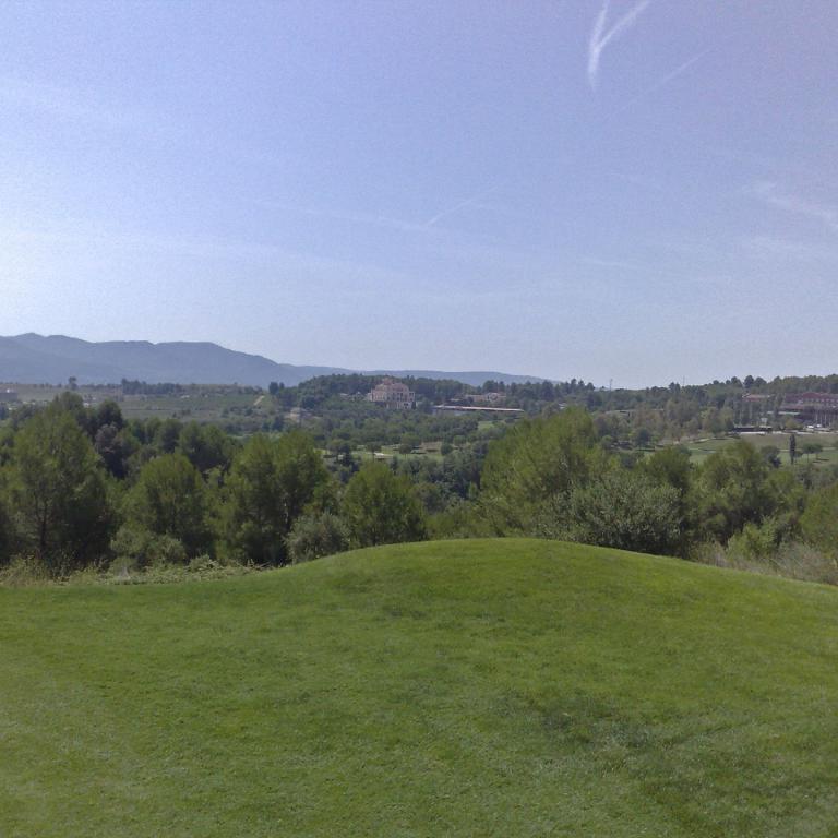 Panoramablick von einem grünen Hügel auf ein darunter liegendes Tal mit Bäumen und Bebauung unter blauem Himmel.