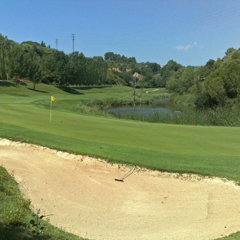 Golfplatz mit Sandbunker im Vordergrund, einer grünen Wiese, einem Teich und dichtem Wald im Hintergrund