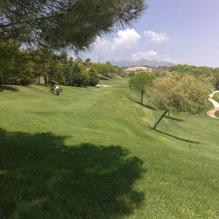 Eine weite, grüne Landschaft, die einen Golfplatz zeigt, mit Bäumen im Vordergrund und einer Bergkette im Hintergrund.