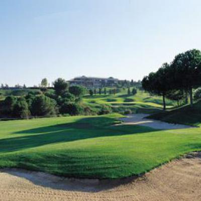 Panoramablick auf einen Golfplatz mit einem großen Sandbunker im Vordergrund, einem grünen Fairway, Bäumen und einem klaren Himmel.
