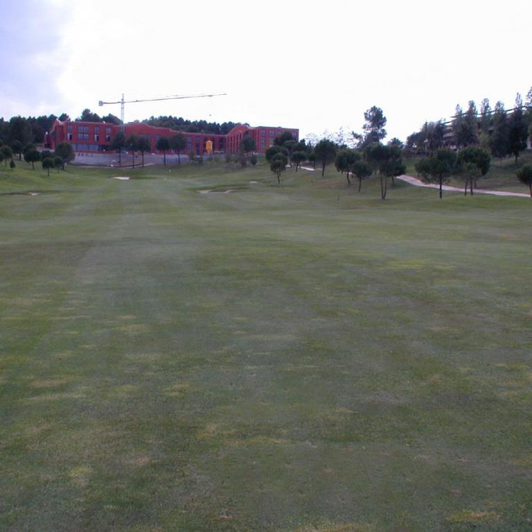 Weitwinkelaufnahme eines großen, gepflegten Golfplatzes mit Gebäuden und Bäumen im Hintergrund.