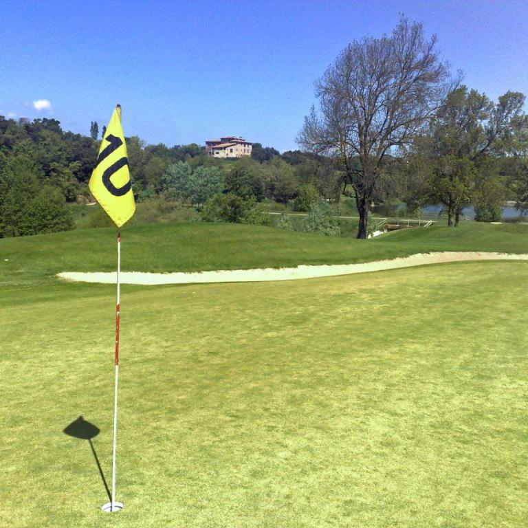 Ein sonniger Blick über einen Golfplatz mit einem Fahnenmast und einem Sandbunker im Vordergrund, dahinter liegt ein Gewässer mit Bäumen und einem Geb