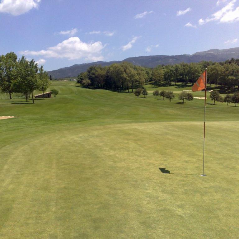 Eine weite Graskoppel eines Golfplatzes mit einem roten Fahnenpfahl und Blick auf bewaldete Berge im Hintergrund.