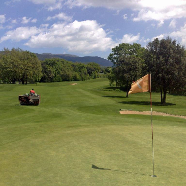Weite Aufnahme eines Golfplatzes mit Fahnenmast und Blick auf bewaldete Hügel.
