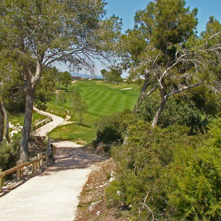 Ein Weg führt durch üppiges Buschwerk und Bäume zu einem großen, gepflegten Golfplatz.