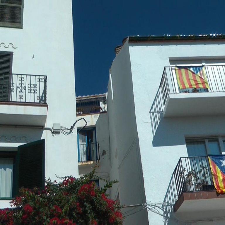 Eine Reihe weißer, mediterraner Wohngebäude mit roten Blumen im Vordergrund und spanischen Flaggen an den Balkonen.