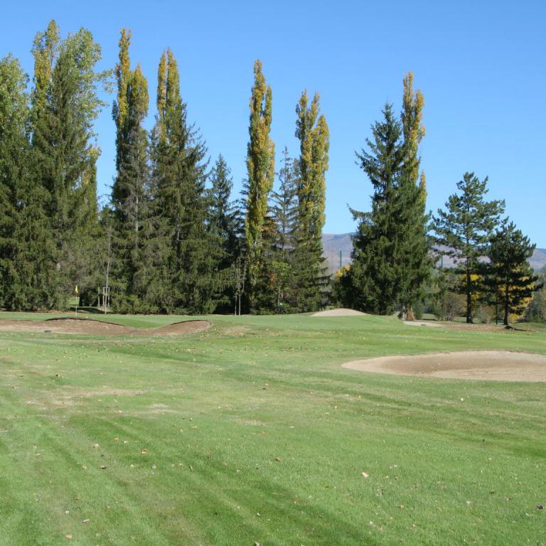 Eine weitläufige Golfplatzlandschaft mit einem Sandbunker im Vordergrund und einer dichten Reihe hoher Bäume im Hintergrund unter blauem Himmel.
