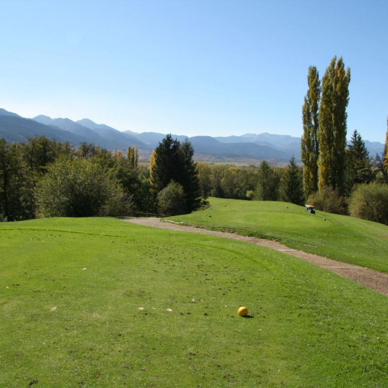 Weitläufiger Blick auf einen Golfplatz mit grünem Rasen, einem Weg und Bergketten im Hintergrund.