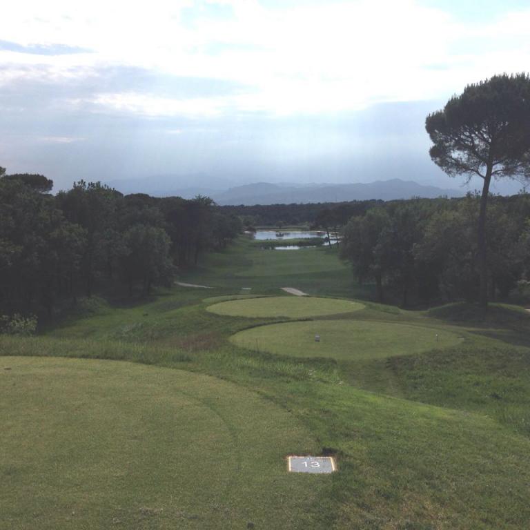 Eine Panoramaaufnahme eines weitläufigen Golfplatzes mit einem Grün im Vordergrund, einem Wasserlauf und Bäumen im Hintergrund.