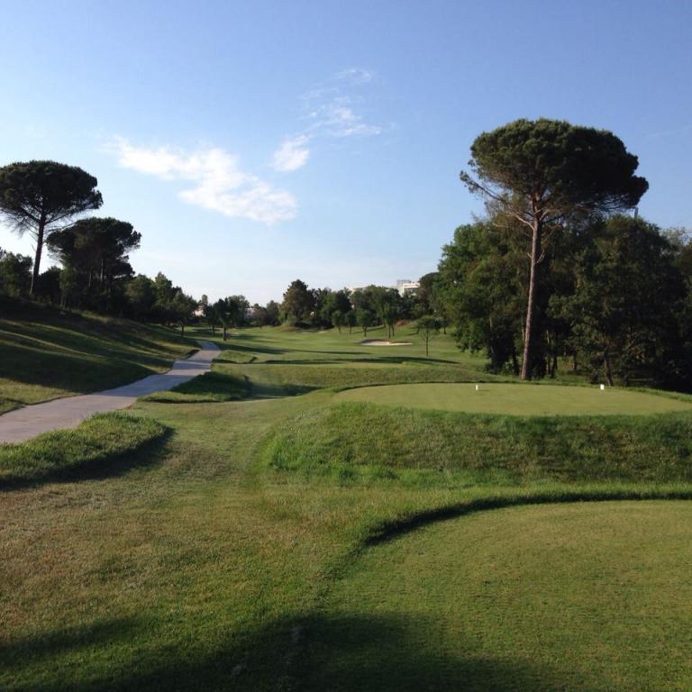 Eine weitläufige Aufnahme eines Golfplatzes mit grünen, gepflegten Rasenflächen, einem gewundenen Weg und zahlreichen Bäumen unter blauem Himmel.