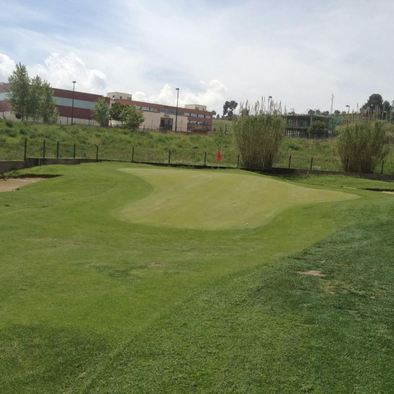 Grüner Golfplatz mit einem Sandbunker und einem dahinter liegenden Gebäude im Hintergrund.