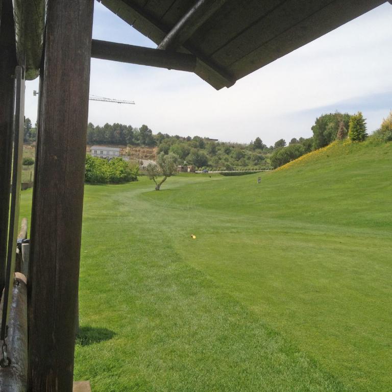 Panoramablick auf einen weitläufigen, gepflegten Rasen, wahrscheinlich ein Golfplatz, aufgenommen von unter einer Holzkonstruktion.