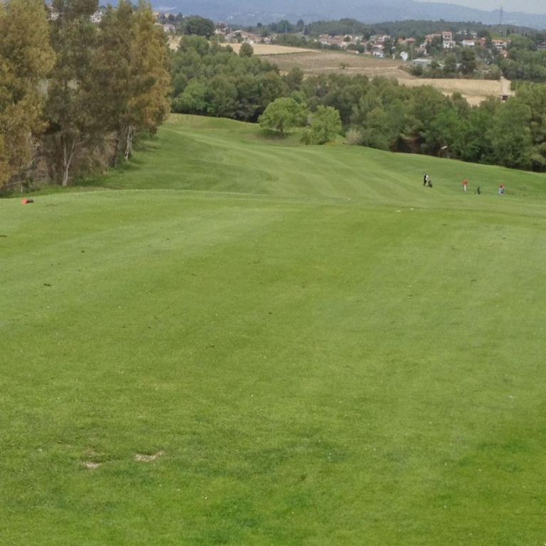 Eine weitläufige Rasenfläche eines Golfplatzes mit Bäumen und einer hügeligen Landschaft im Hintergrund.