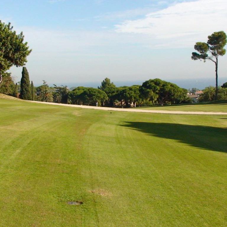 Weitwinkelaufnahme eines Golfplatzes mit Grünfläche im Vordergrund und Bäumen, die einen Blick auf das Meer freigeben.