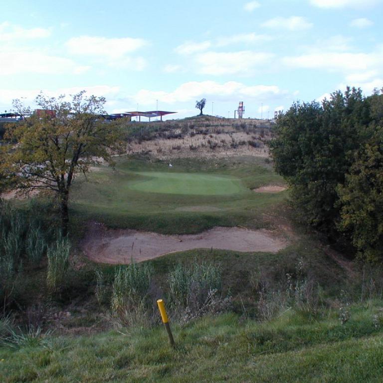 Panoramaaufnahme eines Golfplatzes mit Sandbunker und umgebender Bäumen unter einem bewölkten Himmel.