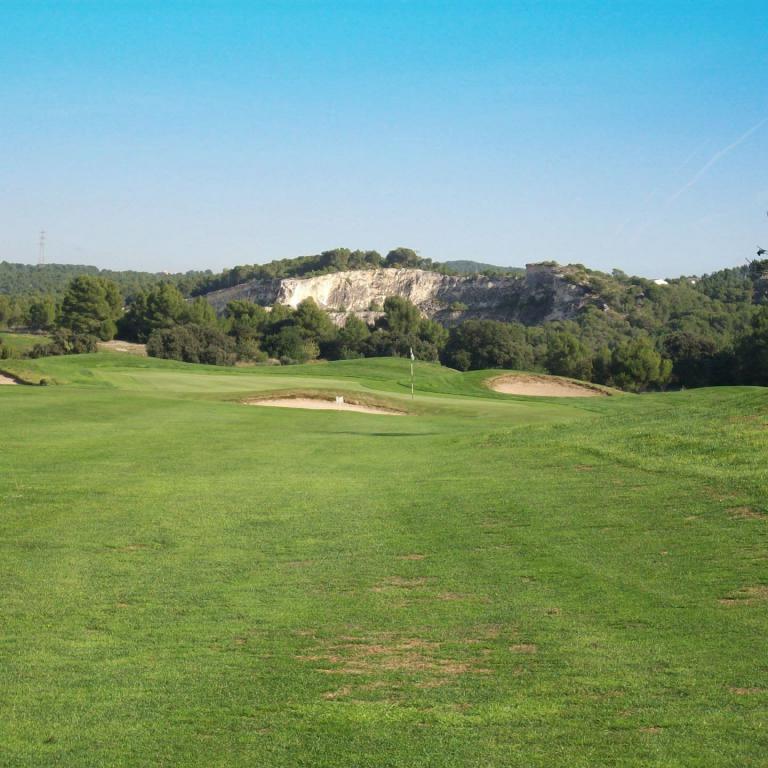 Eine weitläufige Aufnahme eines Golfplatzes mit grünem Rasen im Vordergrund, bewaldeten Hügeln und einer hellen Klippenformation im Hintergrund unter