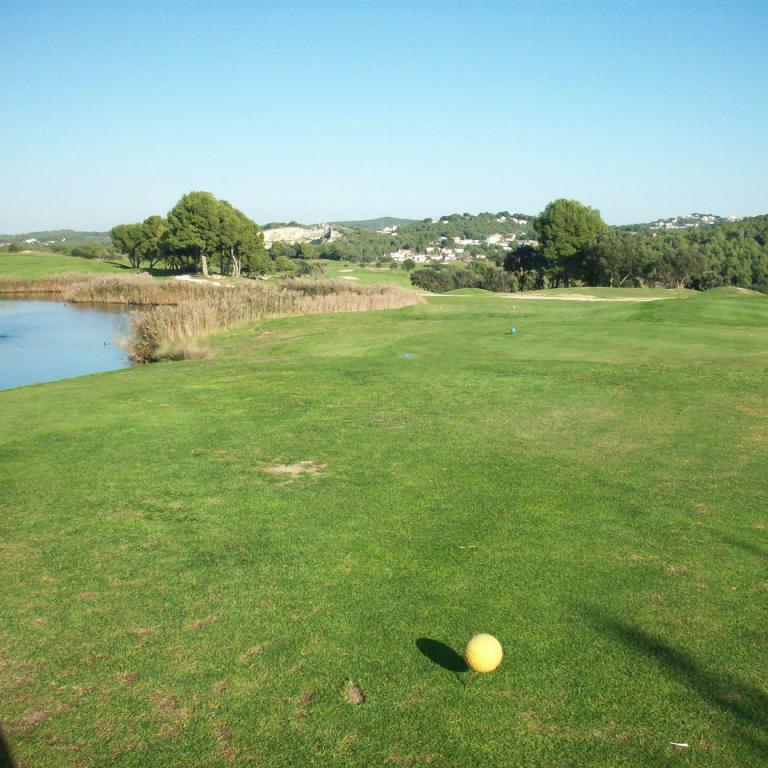 Golfplatz mit einem Golfball im Vordergrund, einem Wasserlauf und bewaldeten Hügeln im Hintergrund.