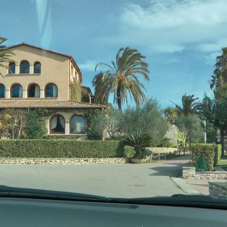 Eine große, mediterrane Villa mit mehreren Fensterreihen, umgeben von Palmen und üppiger Vegetation an einem sonnigen Tag.