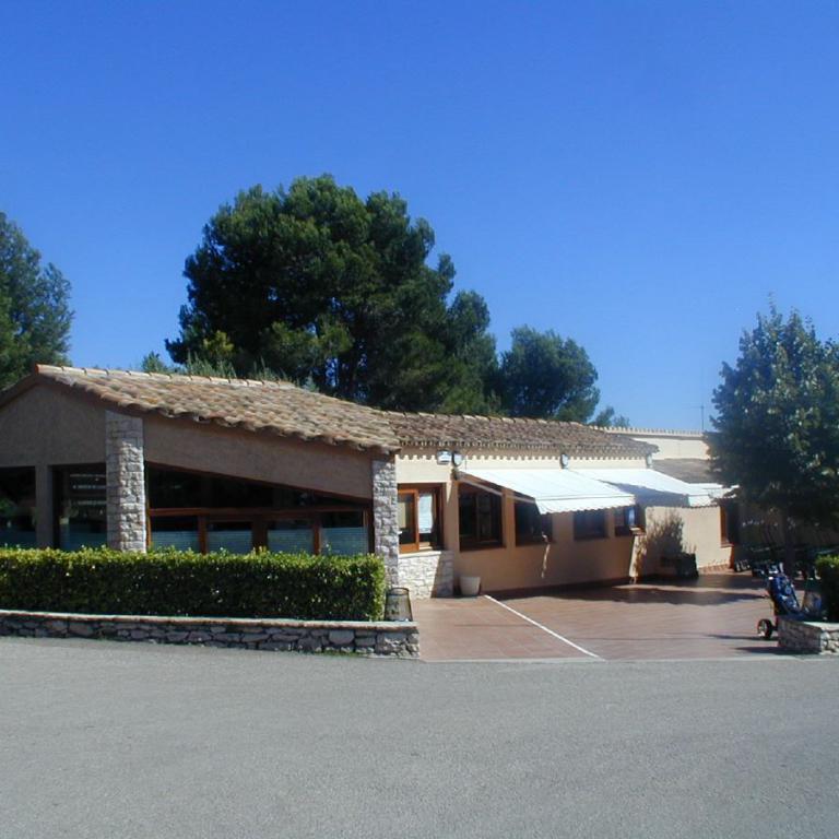 Ein eingeschossiges, mediterranes Gebäude mit Terrakotta-Dach, umgeben von viel Grün und Bäumen unter einem klaren blauen Himmel.