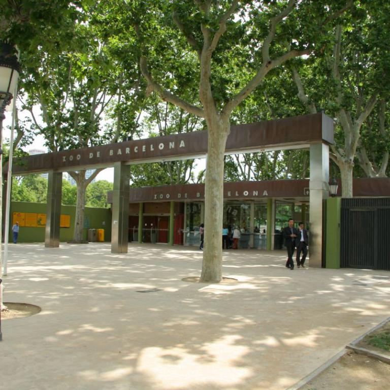 Ein von großen Bäumen umrahmter Platz, der zu einem Eingangsbereich mit der Beschriftung 200 de Barcelona führt.