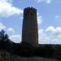 Ein hoher, runder Steinturm steht vor einem blau bewölkten Himmel, umgeben von dichter Vegetation.