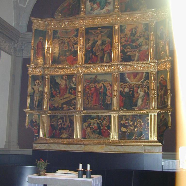 Ein großes, vergoldetes Kirchenretabel mit mehreren Ebenen, das verschiedene biblische Szenen darstellt, vor einem einfachen Tuchtisch.