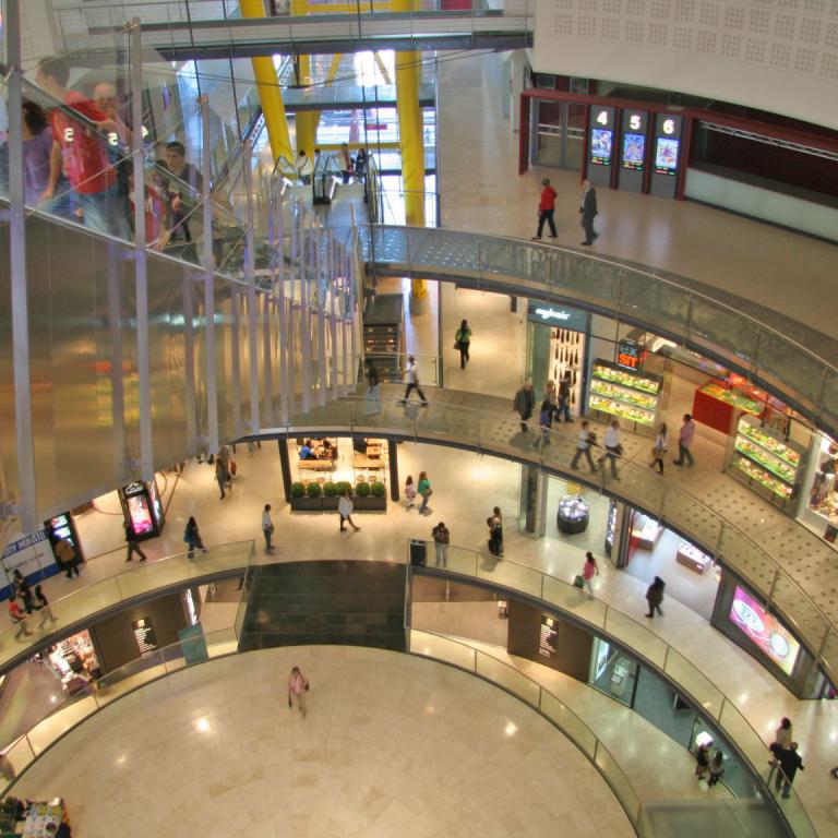 Panoramaaufnahme des weitläufigen Innenraums eines modernen Einkaufszentrums mit mehreren Etagen und Geschäften.