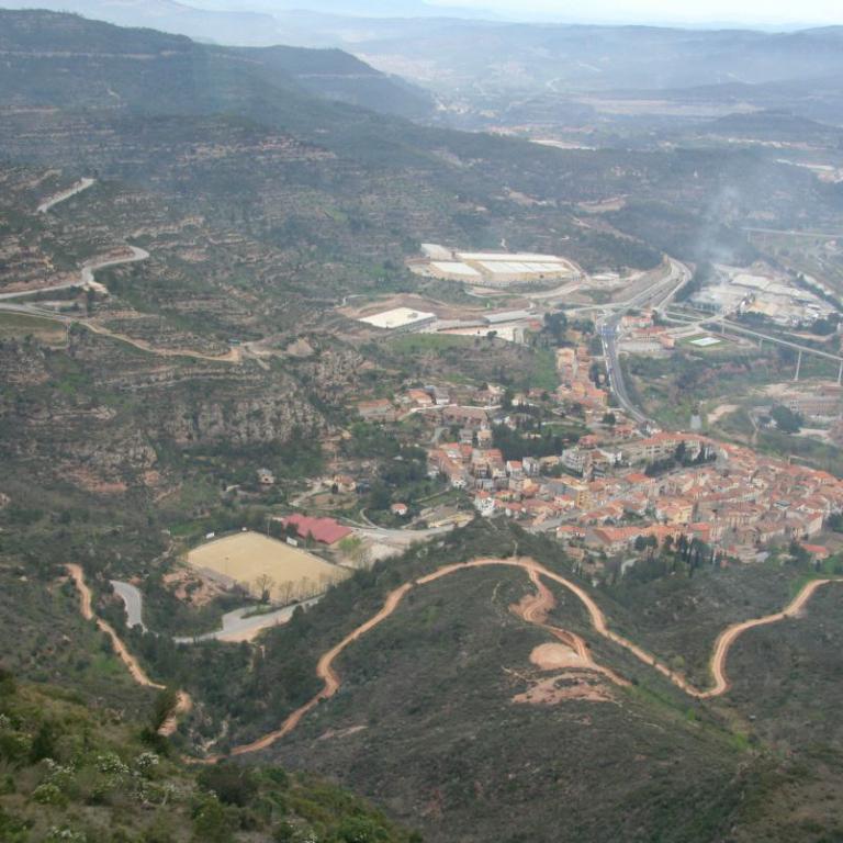 Luftbild eines weitläufigen, hügeligen Gebiets mit erkennbaren Straßen und einer Siedlung im Tal.