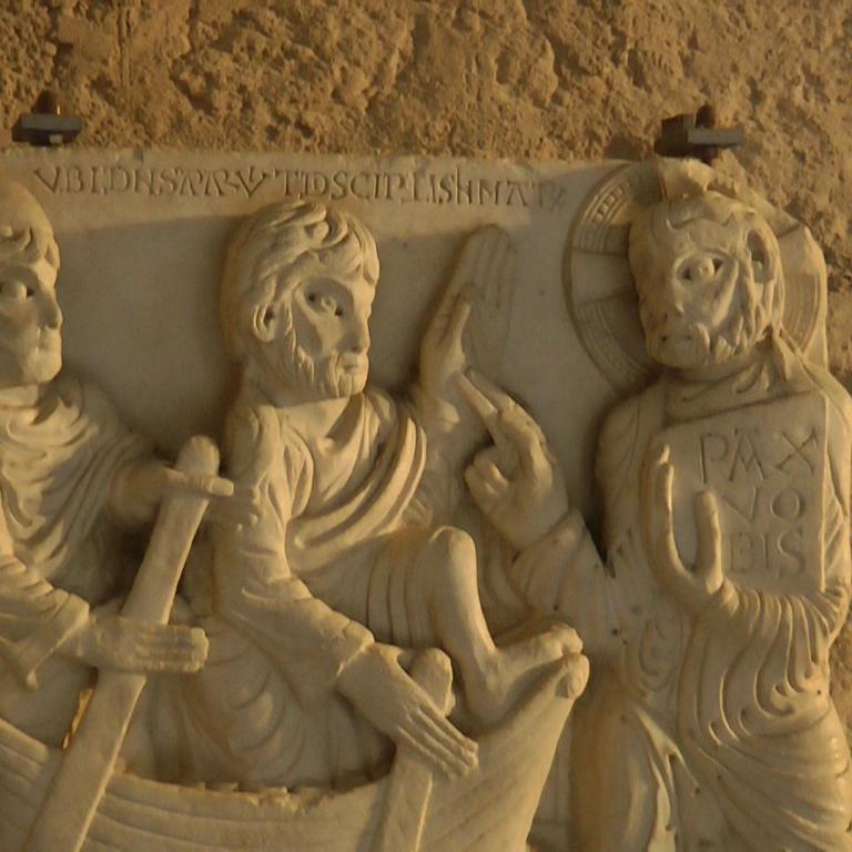 Ein verwittertes Skulpturenrelief, das mehrere Figuren in einem Boot darstellt, eingelassen in eine ockerfarbene Wand.
